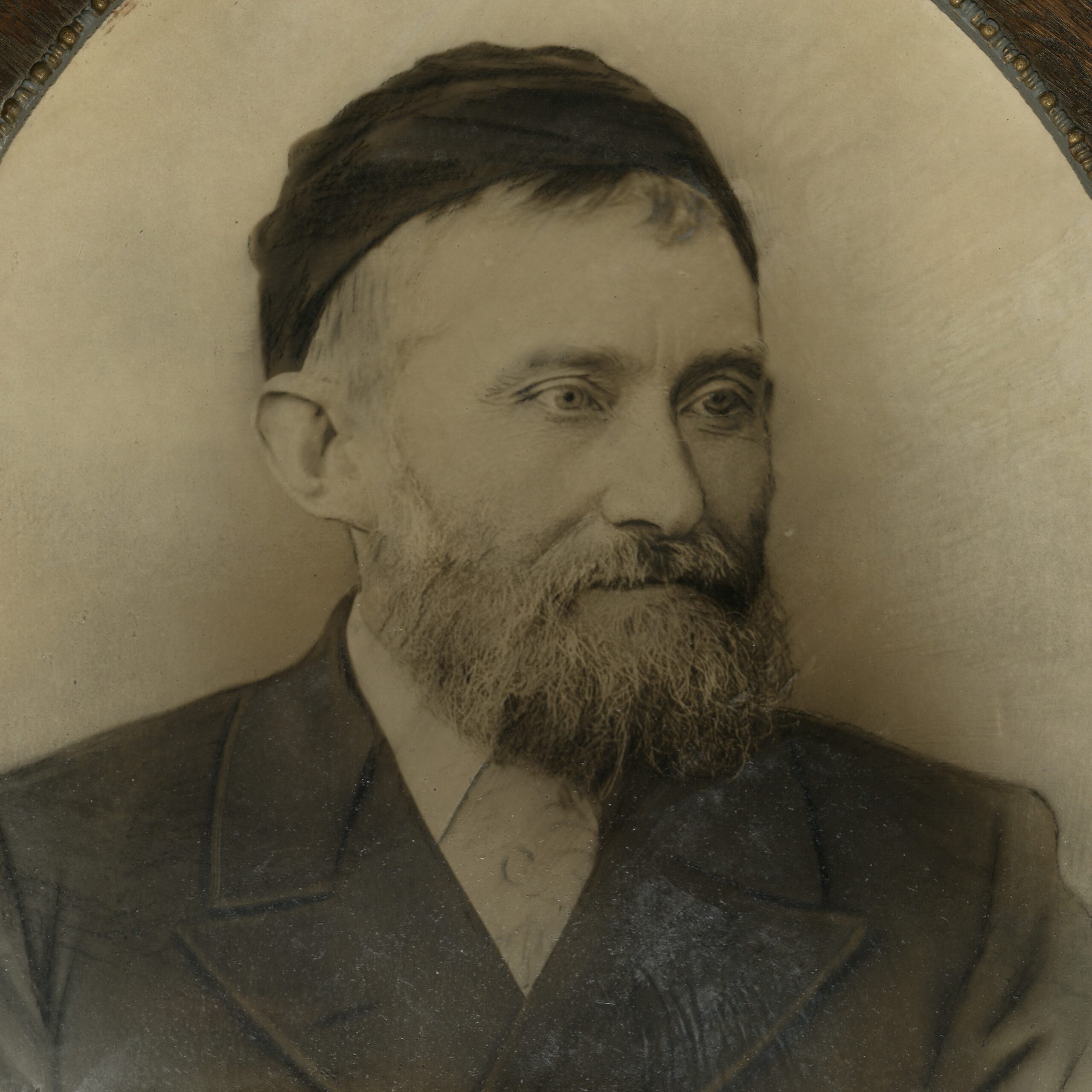 A black and white photo of a man in a dark suit with a full beard and a black cap.